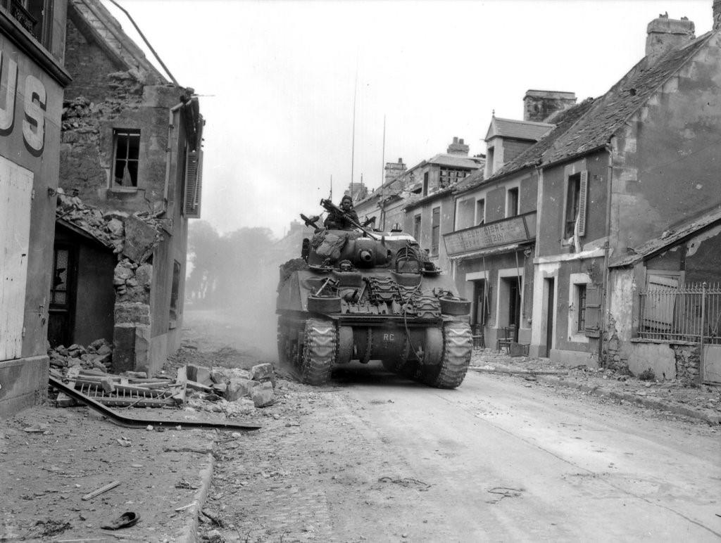 A tank drives through a small village street with crumbling buildings on either side.