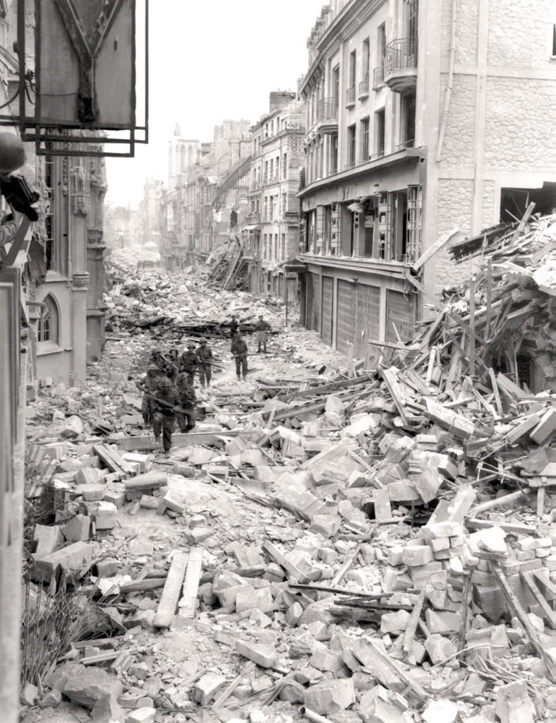 Soldiers walk through a street filled with the wreckage of bombed buildings.