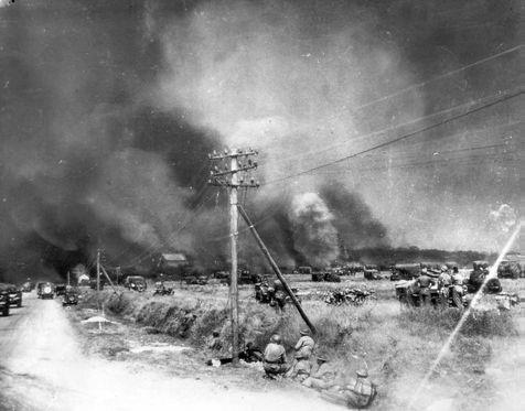Men in a field and under power lines as clouds of smoke fill the air in the distance.
