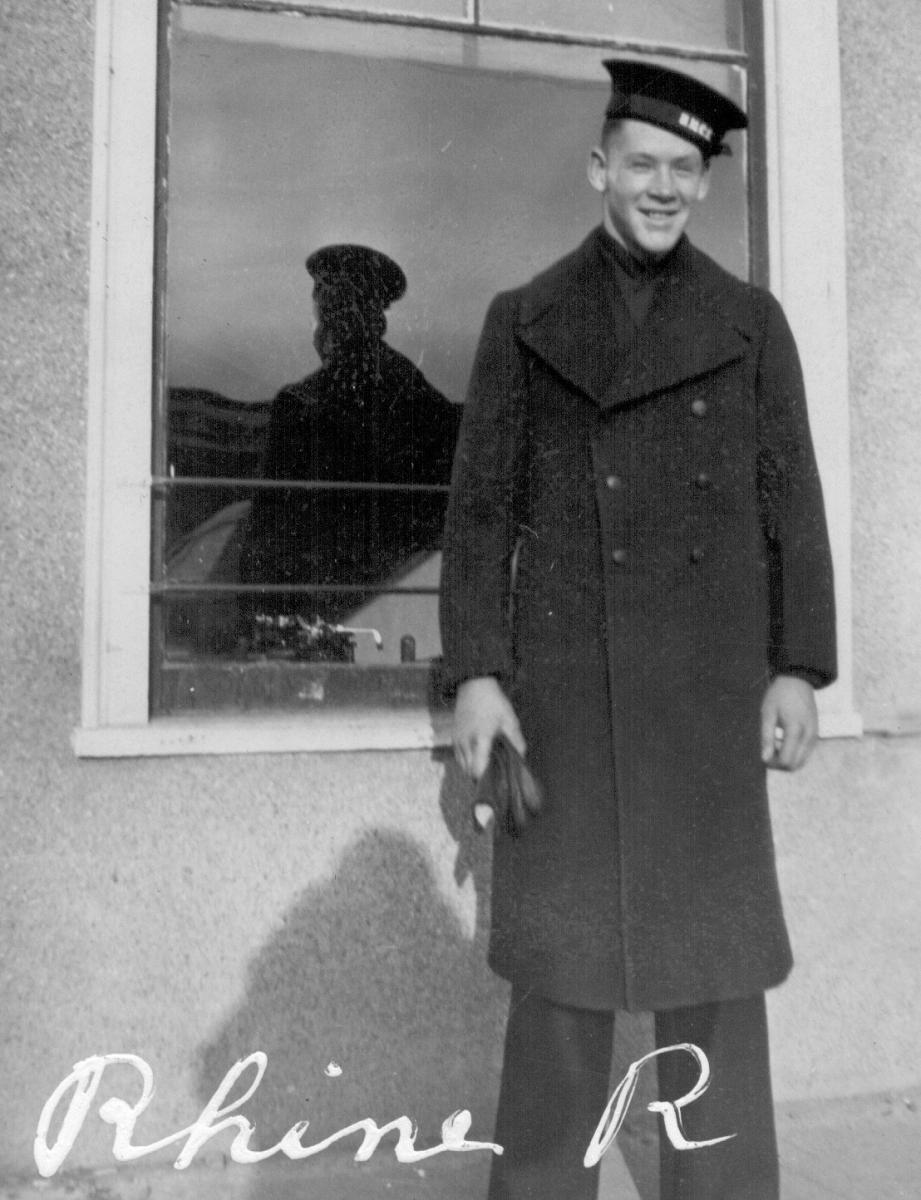 Black and white photo of a man in naval uniform from World War Two standing outside a window.