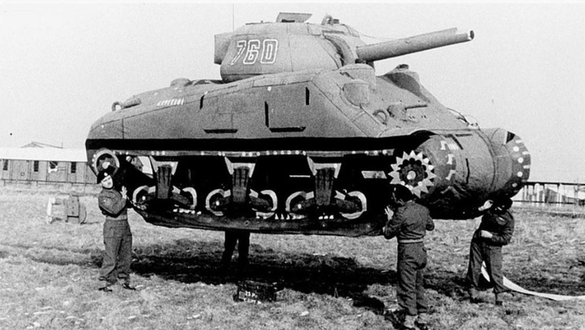 Four men hold an inflatable military tank off the ground.
