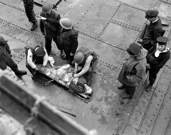 A wounded man lying on the metal deck of a ship, with multiple other men standing around him.