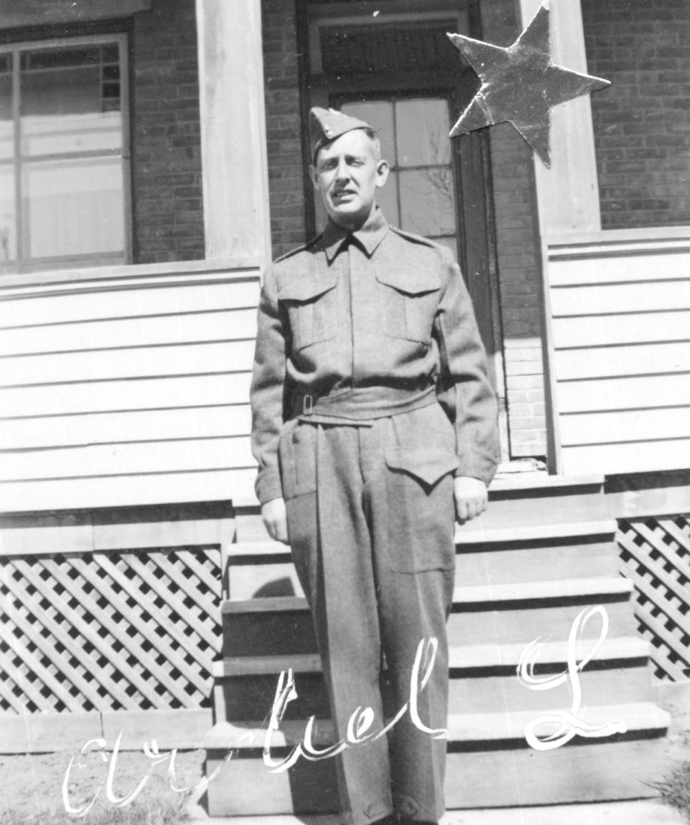 Black and white photo of a man in World War Two army uniform standing by the front steps of a house.