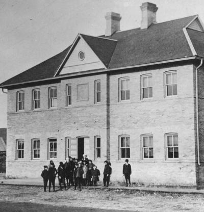 Black and white photograph of the Okotoks Lower School on McRae Street, a large brick building with a group of students coming out the front door.