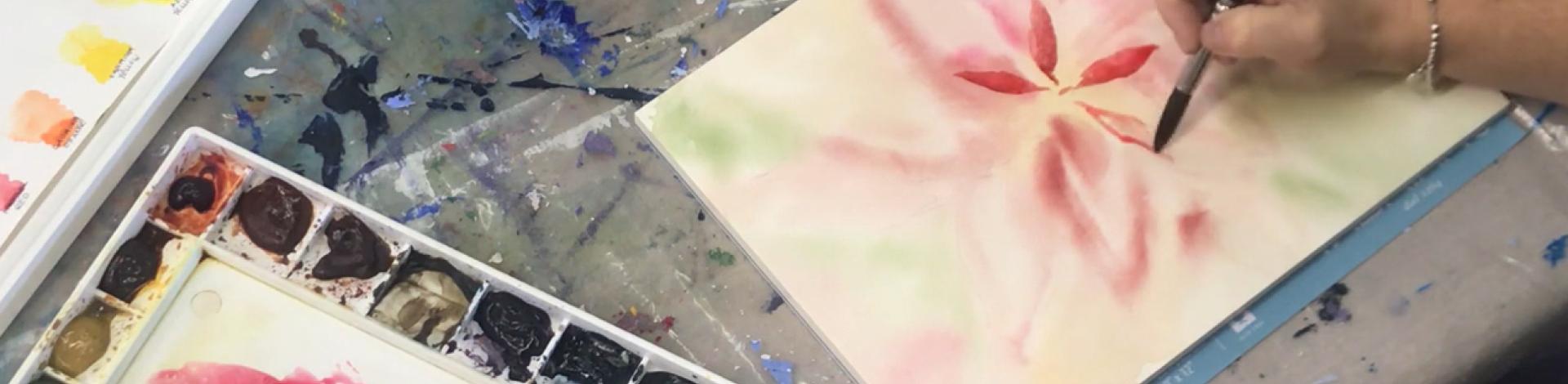 A hand holding a paintbrush, in process of painting a red watercolour flower.