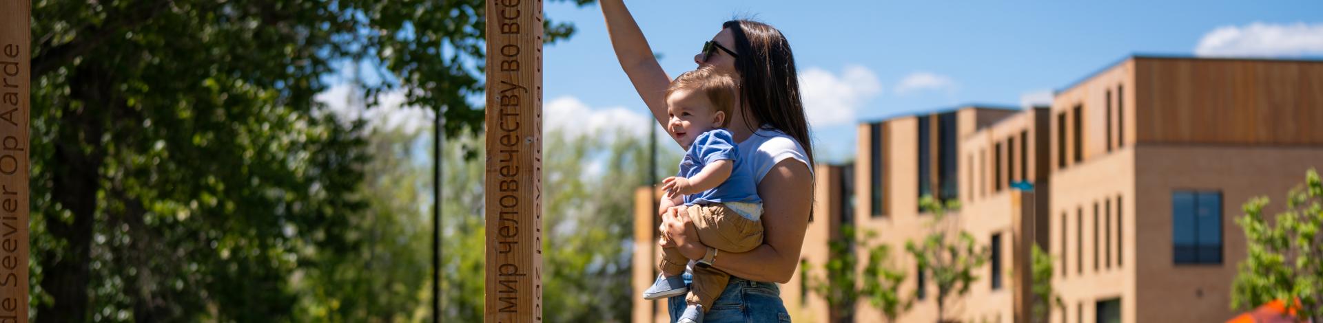 Mother and Baby standing in park touching Peace Poles with Arts & Learning Campus in the background