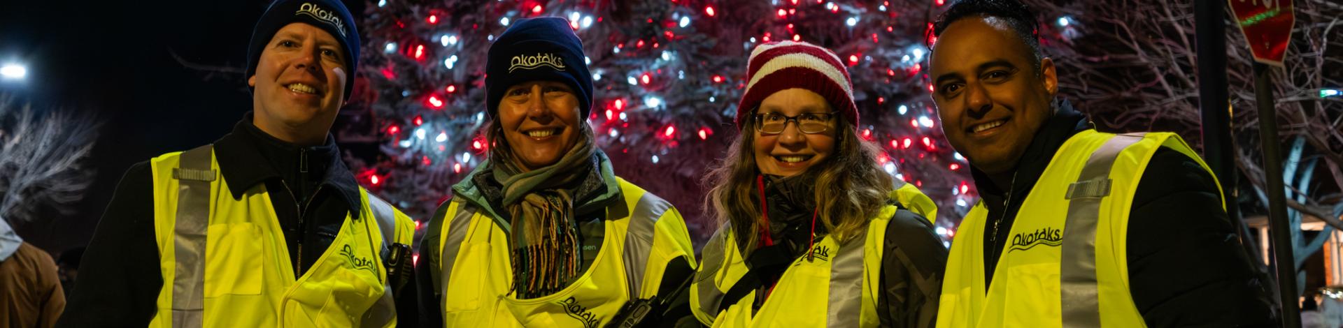 Group of smiling Town staff standing in front of lit Christmas tree while working Light Up Okotoks 2023 