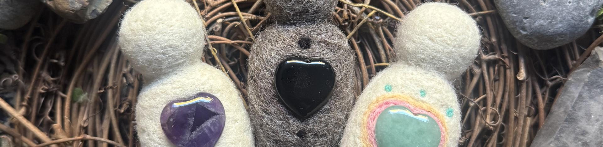 Three felted dolls with hearts made of stones lie in a bird's nest surrounded by rocks. 