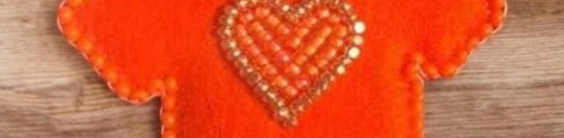 A small pin in the shape of an orange t-shirt, with an orange beaded heart in the centre.