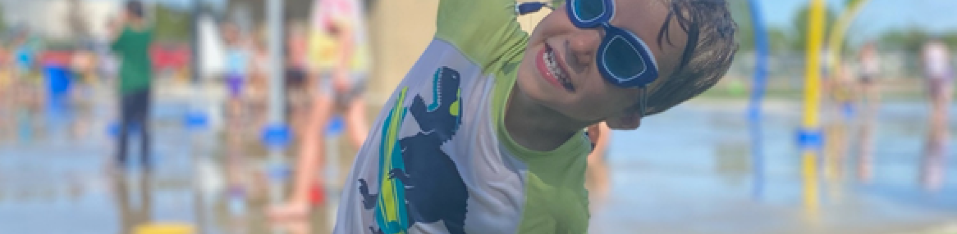 young boy smiling at the spray park with arms outstretched