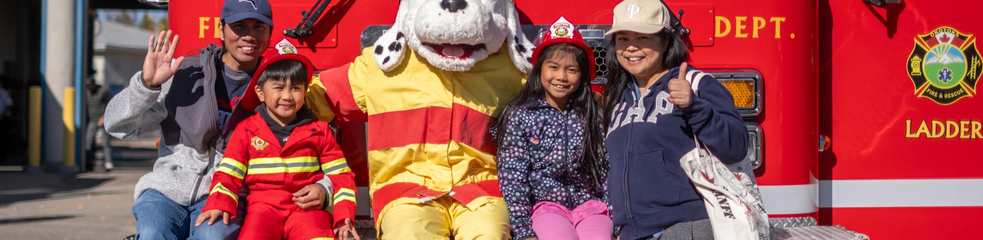 A family of four poses with Sparky in front of a Firetruck