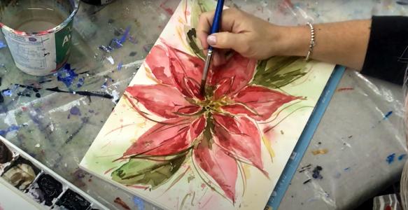A hand painting a red flower with green leaves.