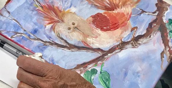The hands of an older person are seen next to an amateur painting of a bird.