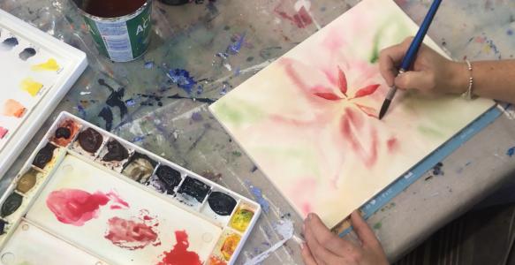 A hand holding a paintbrush, in process of painting a red watercolour flower.