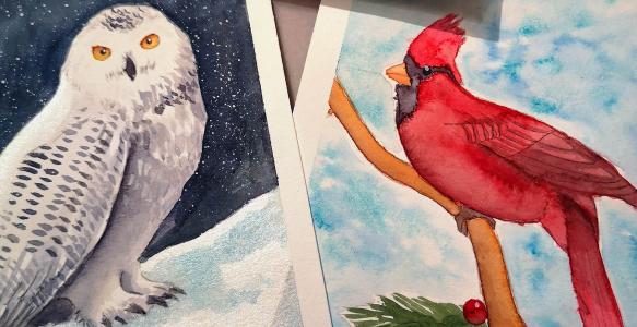 Paintings of a white snowy owl and a red cardinal are lying on a flat surface.