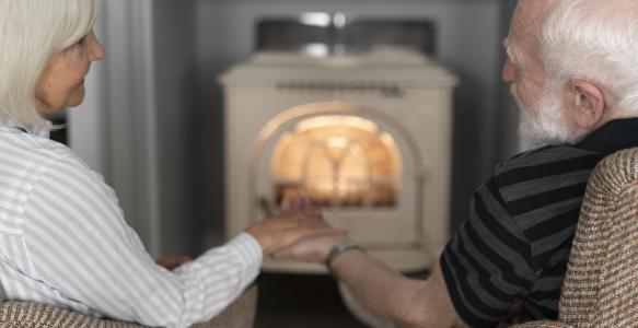 A male and female adult senior sit holding hands looking at each other in front of a fire 
