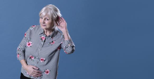 Senior woman in front of blue background putting a hand up to her ear to signal that she is listening 
