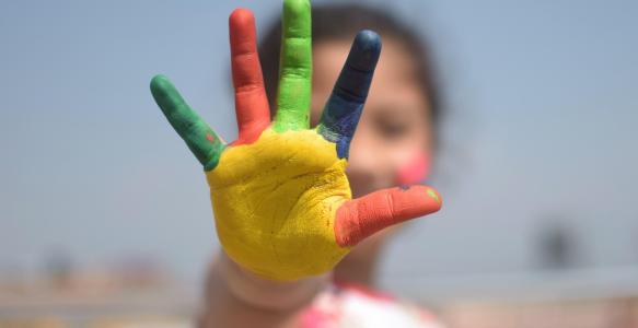 A close-up photo of a young child's hand covered in paint, with each finger a different colour.