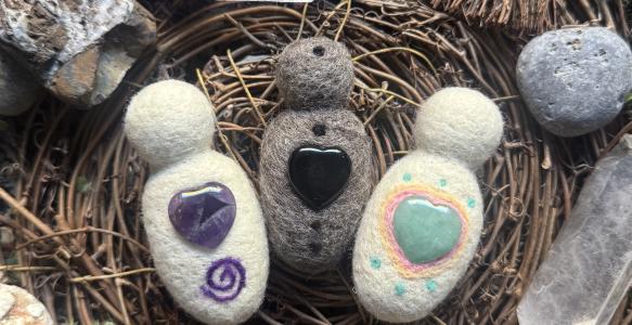 Three felted dolls with hearts made of stones lie in a bird's nest surrounded by rocks. 