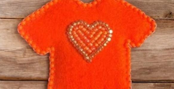 A small pin in the shape of an orange t-shirt, with an orange beaded heart in the centre.