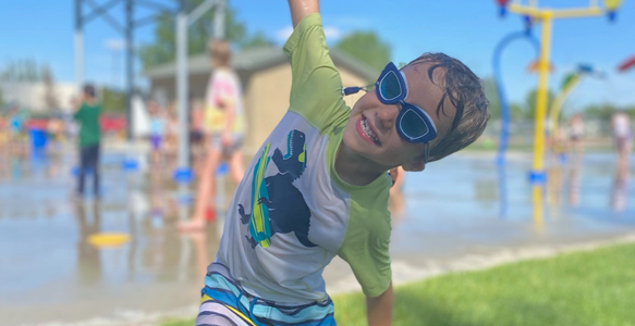 young boy smiling at the spray park with arms outstretched 