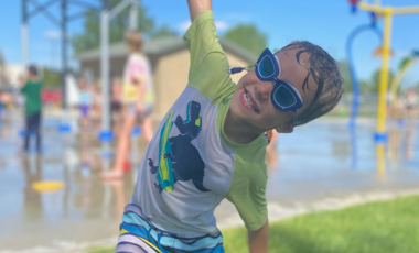 young boy smiling at the spray park with arms outstretched 