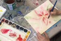A hand holding a paintbrush, in process of painting a red watercolour flower.