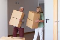 A male senior and female senior both smile as they carry brown moving boxes