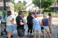 A group of people stand on the sidewalk listening to a woman speak.