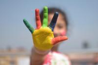 A close-up photo of a young child's hand covered in paint, with each finger a different colour.