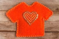 A small pin in the shape of an orange t-shirt, with an orange beaded heart in the centre.