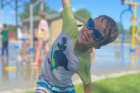 young boy smiling at the spray park with arms outstretched 