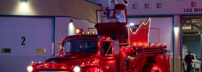 Santa on old fire truck with christmas lights on it in front of fire house