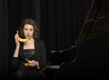 Pianist Sarah Hagan stands in front of a black background holding a banana like a phone.