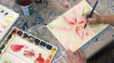 A hand holding a paintbrush, in process of painting a red watercolour flower.