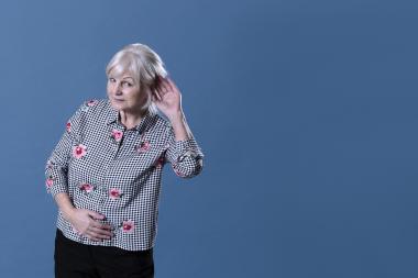 Senior woman in front of blue background putting a hand up to her ear to signal that she is listening 