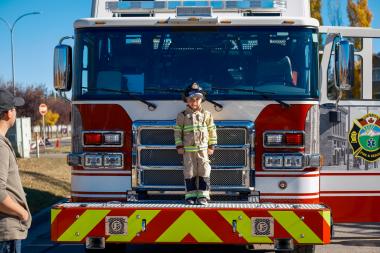 Young boy dressed as fire fighter stands on Okotoks fire engine during Fire Prevention Week